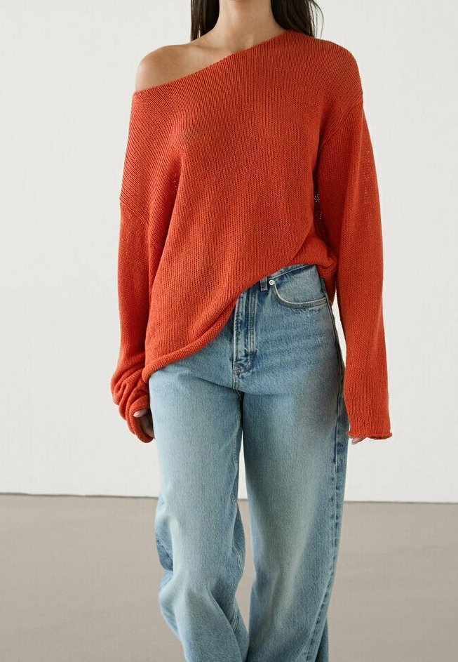 Femme portant un pull en maille orange à épaules dénudées et un jean ample taille haute bleu clair, debout devant un fond uni.