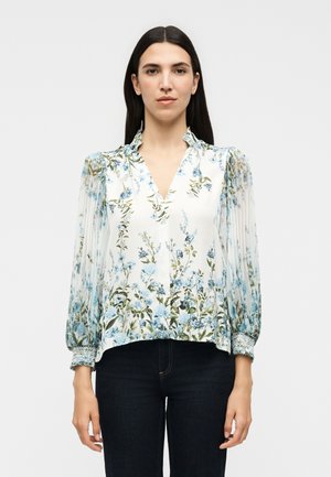 Žena s dlhými tmavými vlasmi oblečená v bielej kvetinovej blúzke s modrými kvetmi a priesvitnými dlhými nahustenými rukávmi, v kombinácii s tmavými nohavicami.