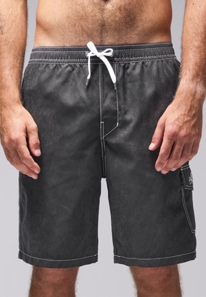 Mann trägt schwarze knielange Badeshorts mit weißem Kordelzug und Seitentasche, steht mit entspannten Armen an den Seiten.