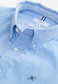 Camisa de botón azul claro hecha de algodón con un acabado texturizado. Presenta un cuello puntiagudo, botones blancos y un logo bordado en el pecho.
