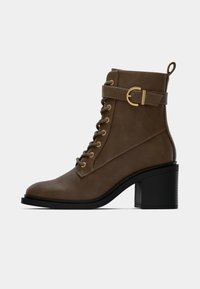 Καφέ δερμάτινο μποτίνι στον αστράγαλο με στρογγυλή μύτη, κορδόνια μπροστά, χρυσά αξεσουάρ και τακούνι τύπου μπλοκ. Διαθέτει λουρί με λεπτομέρεια από αγκράφα.