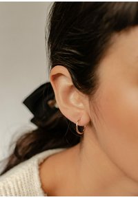 Roségoldene Creolen an einem durchstochenen Ohr, mit einer glatten, glänzenden Oberfläche. Der Hintergrund umfasst ein dunkles Band und einen strukturierten Pullover.