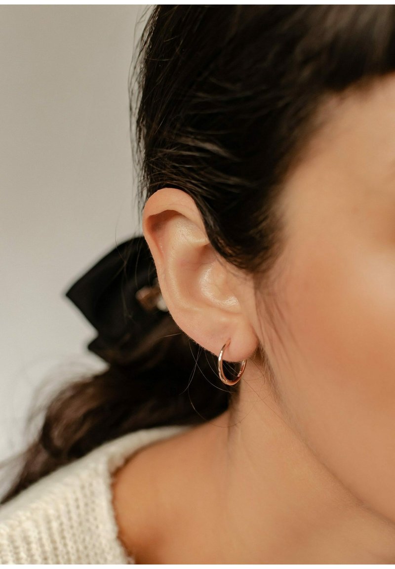 Roségoldene Creolen an einem durchstochenen Ohr, mit einer glatten, glänzenden Oberfläche. Der Hintergrund umfasst ein dunkles Band und einen strukturierten Pullover.