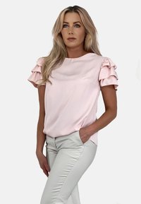 Jasnoróżowa bluzka z falbanami na krótkich rękawach, gładką teksturą, okrągłym dekoltem i luźnym krojem, w zestawieniu z jasnymi dopasowanymi spodniami.