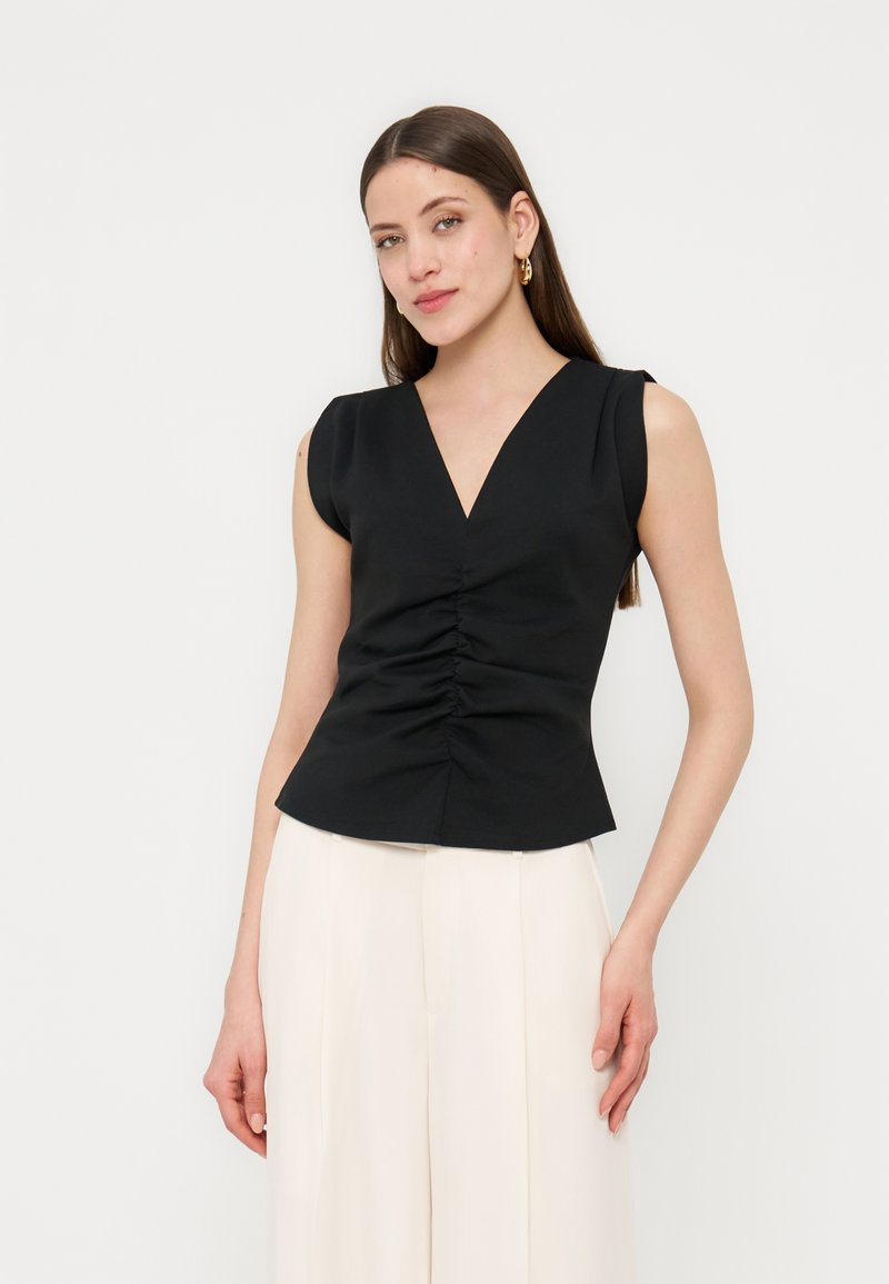 Femme aux longs cheveux bruns portant des boucles d'oreilles créoles dorées, un haut noir sans manches à col en V froncé, et un pantalon blanc taille haute à jambes larges.