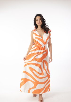 Frau mit langen dunklen Haaren, die ein ärmelloses, orange-weiß gemustertes Maxikleid mit tiefem V-Ausschnitt und Sandalen mit transparenten Riemen trägt.