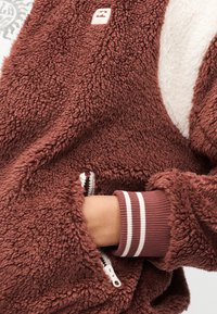 Το χέρι ενός ατόμου βρίσκεται εν μέρει στην τσέπη με φερμουάρ ενός καφέ τζάκετ με χνουδωτή υφή και μανσέτα με λευκές ρίγες στη ριμπ ύφανση του μανικιού.