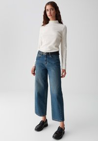 Cremfarbenes Langarmshirt mit subtilen Sprenkeln, kombiniert mit hochgeschnittenen blauen weit geschnittenen Jeans und schwarzen Schnürschuhen. Einfaches und modernes Design.
