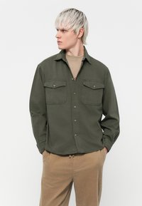 Olive green button-up shirt with two chest pockets, collar, and long sleeves. Worn over a beige crew-neck shirt and paired with tan trousers.