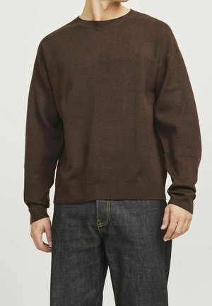 Mann trägt einen schlichten braunen Pullover mit Rundhalsausschnitt und langen Ärmeln sowie dunkelblaue Jeans, steht vor einem einfarbigen hellen Hintergrund.