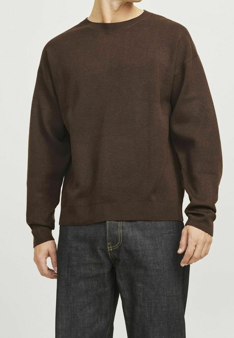 Homme portant un pull à col rond marron uni à manches longues et un jean bleu foncé, debout devant un fond clair uni.