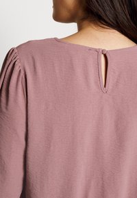 Blouse rose avec des manches ballon, une ouverture en forme de cœur à l'arrière et un tissu doux et texturé. Design simple et élégant.