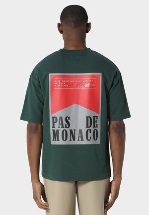 Homme portant un T-shirt vert foncé avec un grand graphisme rouge et gris dans le dos affichant le texte "PAS DE MONACO" et un texte plus petit au-dessus.