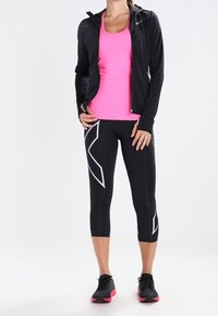 Schwarze Sportjacke über einem pinken Tanktop, kombiniert mit schwarzen, knöchellangen Leggings mit weißen geometrischen Mustern und pinken Akzent-Schuhen.