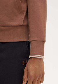 Felpa marrone con polsi a coste, caratterizzata da strisce bianche a contrasto. Il polsino presenta una texture liscia, abbinata a pantaloni scuri e un logo ricamato.