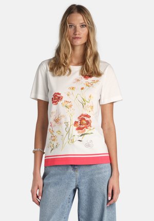 Femeie blondă care poartă un tricou alb cu imprimeu floral roz și roșu și tiv coral, asortat cu blugi albastru deschis.