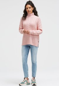 Rosa gestrickter Pullover mit hohem Kragen, strukturiertem Muster und seitlichen Schlitzen, kombiniert mit hellblauen Jeans und mehrfarbigen Sportschuhen.