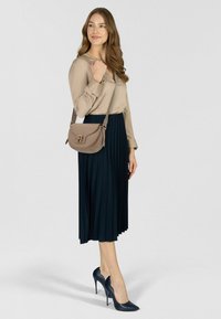Donna in piedi che indossa una camicetta beige, una gonna midi nera plissettata, tacchi alti neri e una borsa a tracolla beige con dettaglio di chiusura.