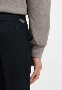 Σκούρο μπλε παντελόνι με πίσω τσέπη και κλείσιμο με κουμπί, με διακριτικό μεταλλικό σύμβολο φτερού. Ορατό γκρι μακρυμάνικο πουκάμισο.