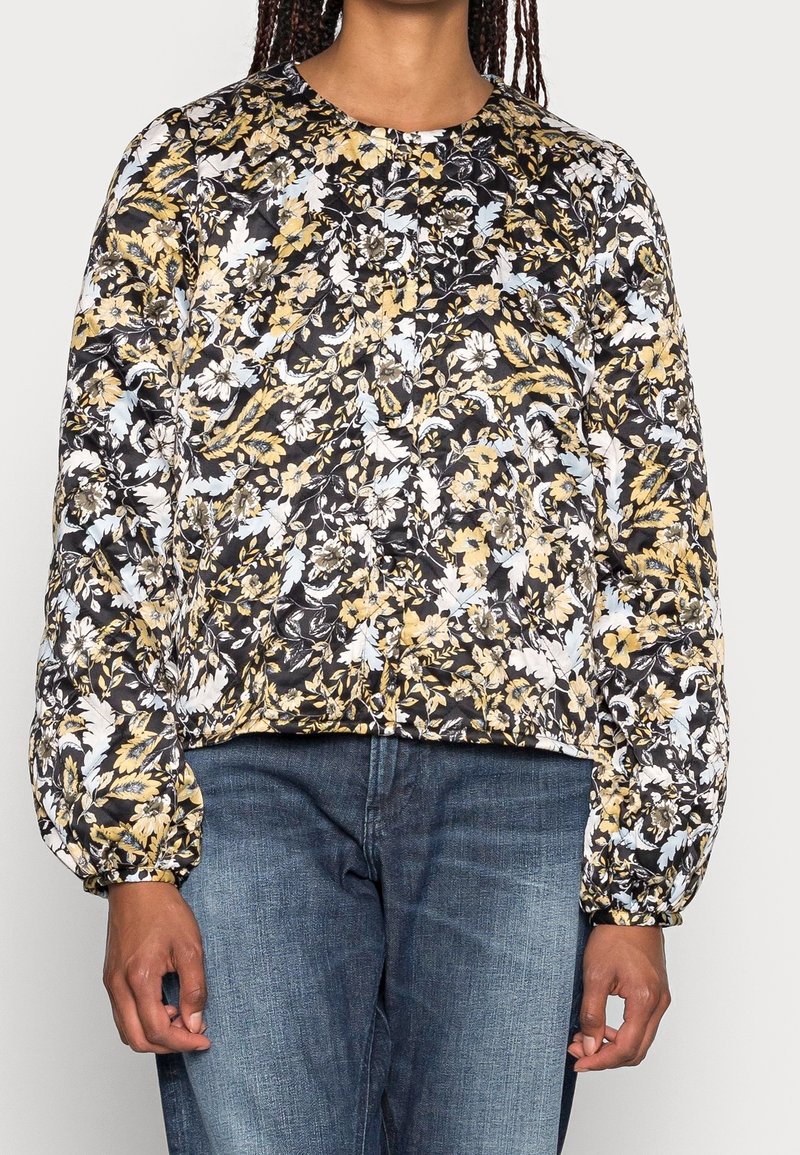 Femme portant un chemisier à manches longues noir à motifs floraux jaunes et blancs, associé à un jean en denim bleu.