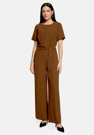 Kvinde iført en brun kortærmet jumpsuit med taljebånd og bukser med vide ben, kombineret med sorte spidse hæle, stående med fronten mod kameraet.