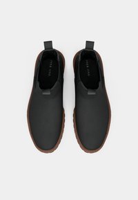 Melni slip-on apavi ar teksturētu virsmu, brūnām gumijas zolēm un cilpām aizmugurē ērtai uzvilkšanai. Minimālistisks dizains.