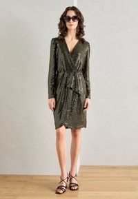 Robe portefeuille à manches longues en tissu métallis�é doré avec des sequins, présentant un décolleté en v, une taille à nouer et une longueur mi-genou. Associée à des sandales noires.