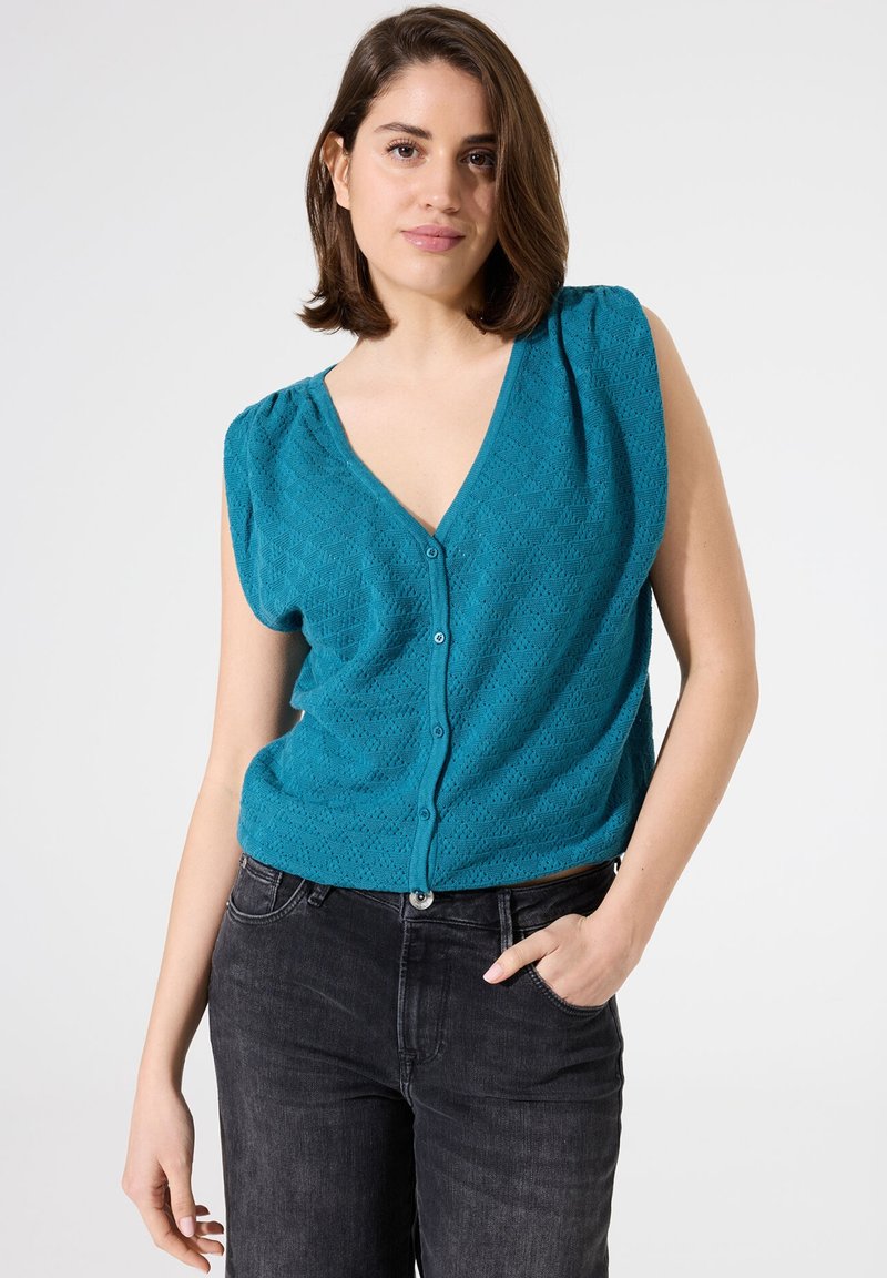 Cárdigan sin mangas en color turquesa con escote en V, patrón texturizado y cierre de botones en la parte delantera, combinado con jeans negros.