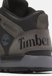 Zapatilla alta gris y negra con material Gore-Tex. Presenta la marca Timberland y el logotipo del árbol. Superficie texturizada y construcción resistente.