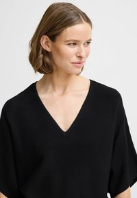 Top en tricot noir avec un col en V, coupe décontractée, manches courtes et texture lisse. Il présente des lignes épurées et un détail minimal.