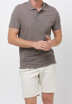 Homme portant un polo marron à manches courtes et un short blanc jusqu'aux genoux, debout, les mains jointes.
