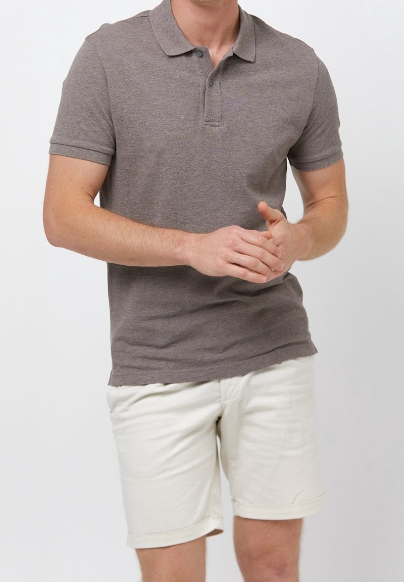 Homme portant un polo marron à manches courtes et un short blanc jusqu'aux genoux, debout, les mains jointes.