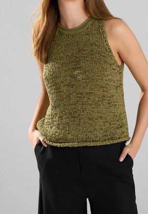 Femme portant un top en tricot vert olive sans manches et un pantalon noir, les mains dans les poches, sur un fond gris uni.