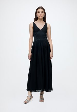 Donna in piedi che indossa un abito lungo nero senza maniche con scollo a V e sandali argentati con cinturini e tacco su sfondo bianco uniforme.