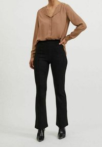 Blusa marrone a maniche lunghe con scollo a V e bottoni frontali, abbinata a pantaloni neri a vita alta. Stivaletti neri con tacco completano l'outfit.