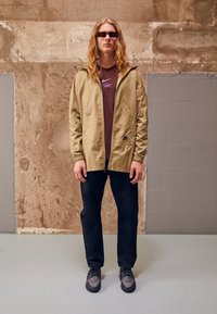Lahka zadrgo jakna v bež barvi z elastičnimi manšeti, v kombinaciji z rjavim T-shirtom. Temni hlače in sivi športni čevlji z črnimi detajli dopolnjujejo obleko.