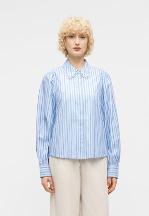 Ξανθός άτομο με σγουρά μαλλιά που φοράει ανοιχτό μπλε ριγωτό μπλούζα με φουσκωτά μανίκια και γιακά Πίτερ Παν με κυματιστό περίγραμμα, καθώς και μπεζ παντελόνι.