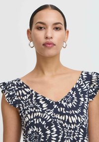 Námornícke šaty s bielym kvetinovým vzorom, výstrih do V a volánovými rukávmi. Modelka nosí kruhové náušnice, minimálny make-up a neutrálny pozadí.