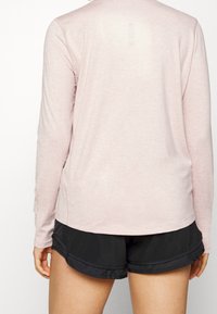 Světle růžový top s dlouhým rukávem vyrobený z měkké tkaniny, s kulatým lemem a malým designovým detailem u výstřihu. Nosí se s černými šortkami.