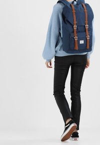 Granatowy plecak z brązowymi skórzanymi paskami i klamrami, wykonany z trwałego materiału. Posiada zapięcie na klapę oraz kieszeń z przodu.