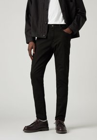 Bărbat îmbrăcat cu blugi negri, pantofi din piele maro închis, șosete albe, cămașă albă și o jachetă neagră, cu o mână în buzunar.