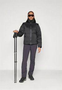 Czarna kurtka puchowa zapinana na zamek, noszona z ciemnymi spodniami i solidnymi czarnymi butami. Użytkownik trzyma kijki trekkingowe; całość o nowoczesnym, eleganckim designie.