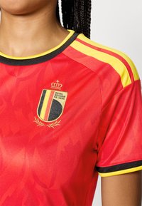 Κόκκιν�η αθλητική φανέλα με κίτρινο και μαύρο φινίρισμα, που φέρει το έμβλημα της Βασιλικής Ποδοσφαιρικής Ομοσπονδίας του Βελγίου και κίτρινες ρίγες στους ώμους.