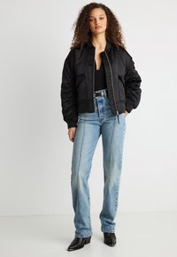 Γυναίκα με σγουρά μαλλιά, φορώντας μαύρο μπουφάν τύπου bomber, μαύρο τοπ, ανοιχτό μπλε τζιν με ψηλή μέση, μαύρη ζώνη και μαύρες μυτερές μπότες, στέκεται όρθια.