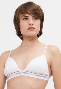 Bralette triangular blanco con tirantes finos, que presenta una banda de marca en gris y rojo. Tejido suave con una textura agradable.