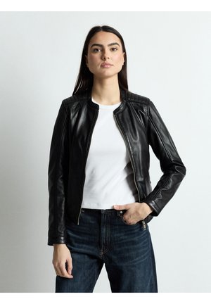 Vrouw met lang donker haar, gekleed in een zwarte leren jas, witte blouse en donkere spijkerbroek, staand tegen een eenvoudige lichte achtergrond.