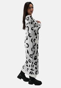 Langes Strickkleid in Weiß mit schwarzem Leopardenmuster. Hat einen lockeren Schnitt, lange Ärmel und seitliche Taschen. Wird mit schwarzen Stiefeln getragen.