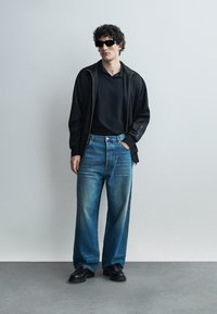 Muž v čiernej bunde na zips, čiernom tričku, modrých džínsach s širokými nohavicami, čiernych topánkach a čiernych slnečných okuliaroch stojí opretý o sivú stenu.