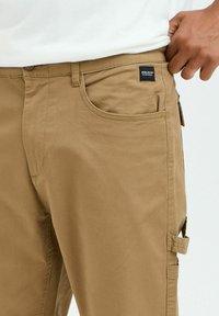 Tan cargo pants made of woven fabric, featuring a front pocket, side pocket, and sturdy stitching. Logo tag on the waistband.