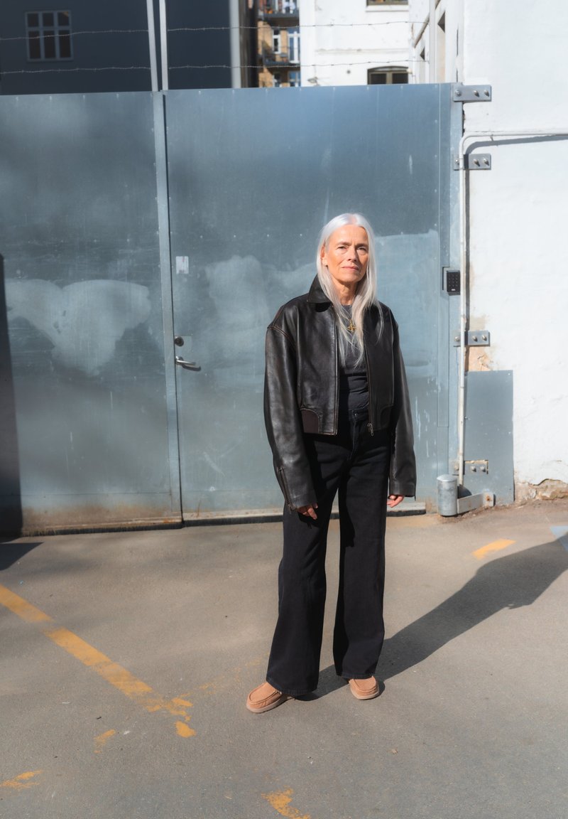 Femme âgée aux longs cheveux gris, portant une veste en cuir noire et un pantalon large noir, debout sur une surface en béton avec une porte métallique derrière elle.